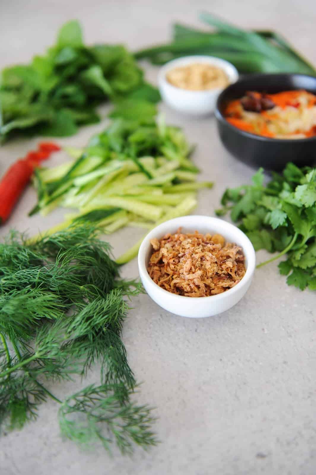 close up of fried shallots in a white bowl, surrounded by herbs and other ingredients