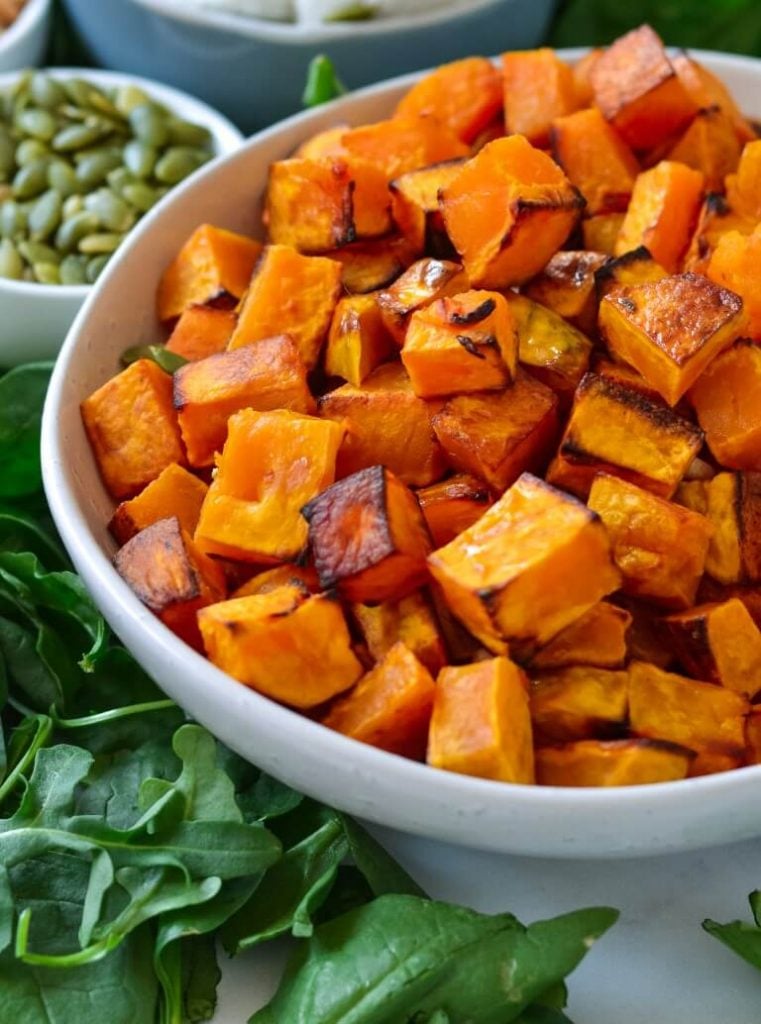 white bowl containing roast pumpkin, surrounded by pine nuts and rocket