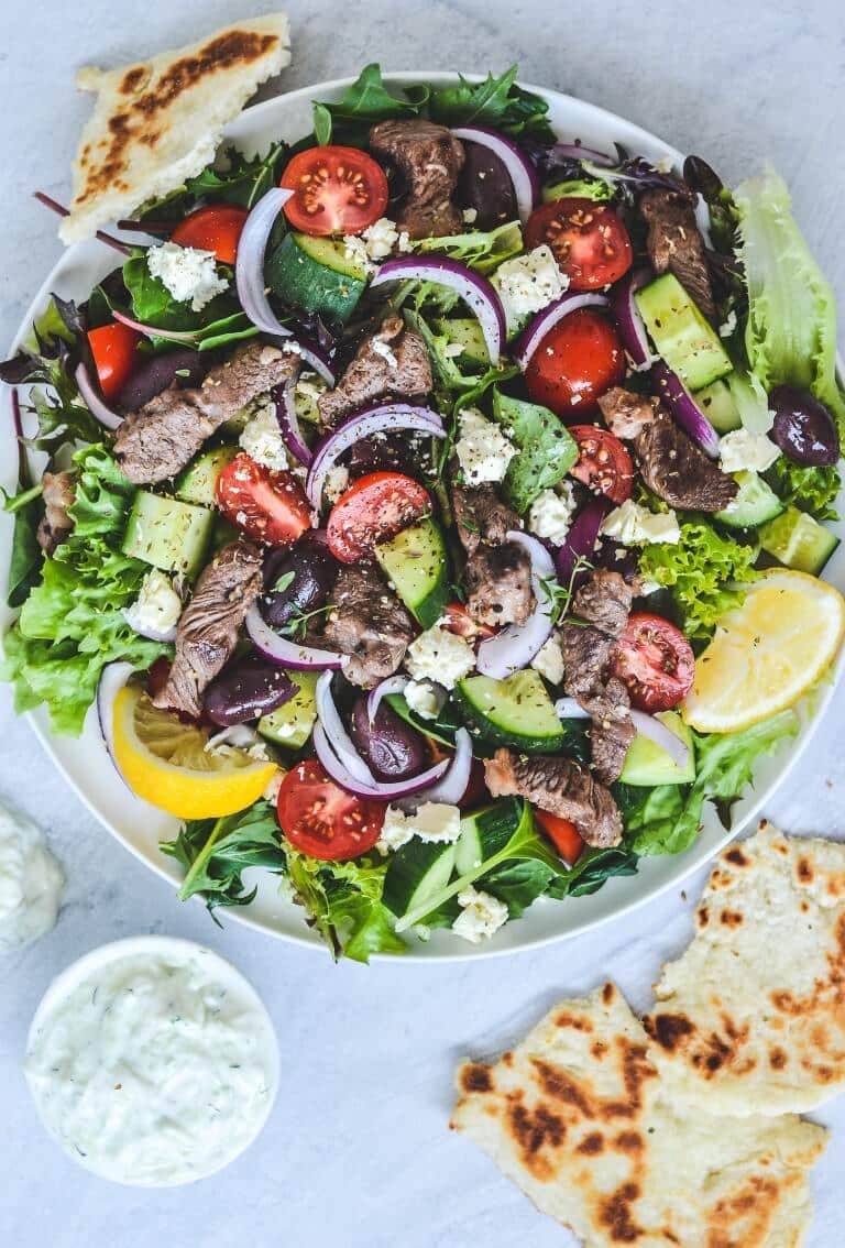 A plate of salad with a slice of flat bread, with a dish of tzatziki