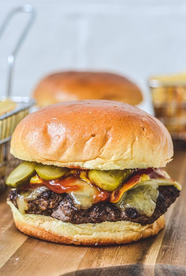 A close up of a burger with pickles, sauce and cheese on wooden board with fries