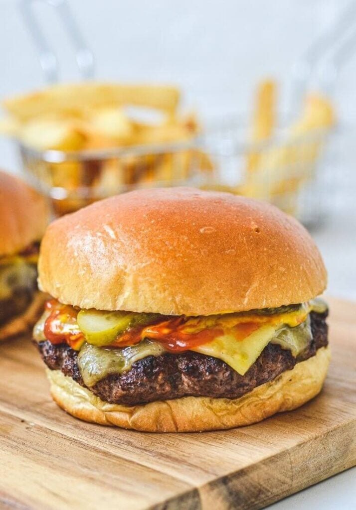 a close up of a cheeseburger on a wooden board with fries in background