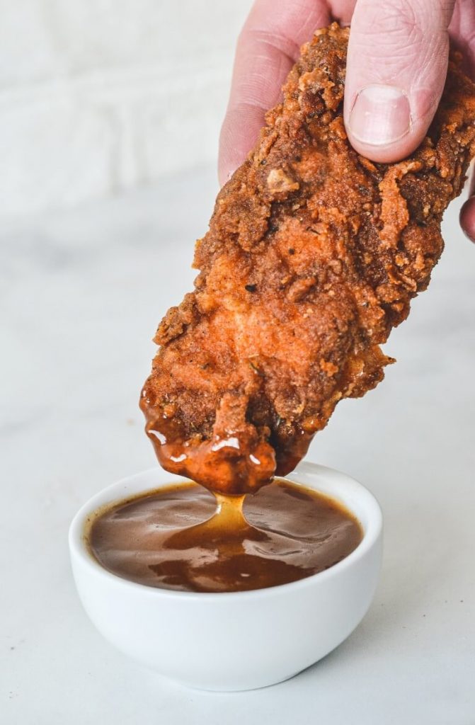 a hand holding a piece of fried chicken, dipping into sauce from white bowl