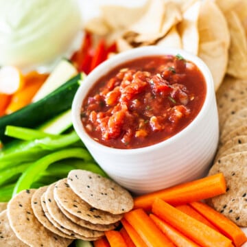 close up of a white bowl with salsa, surrounded by sliced vegetables and crackers