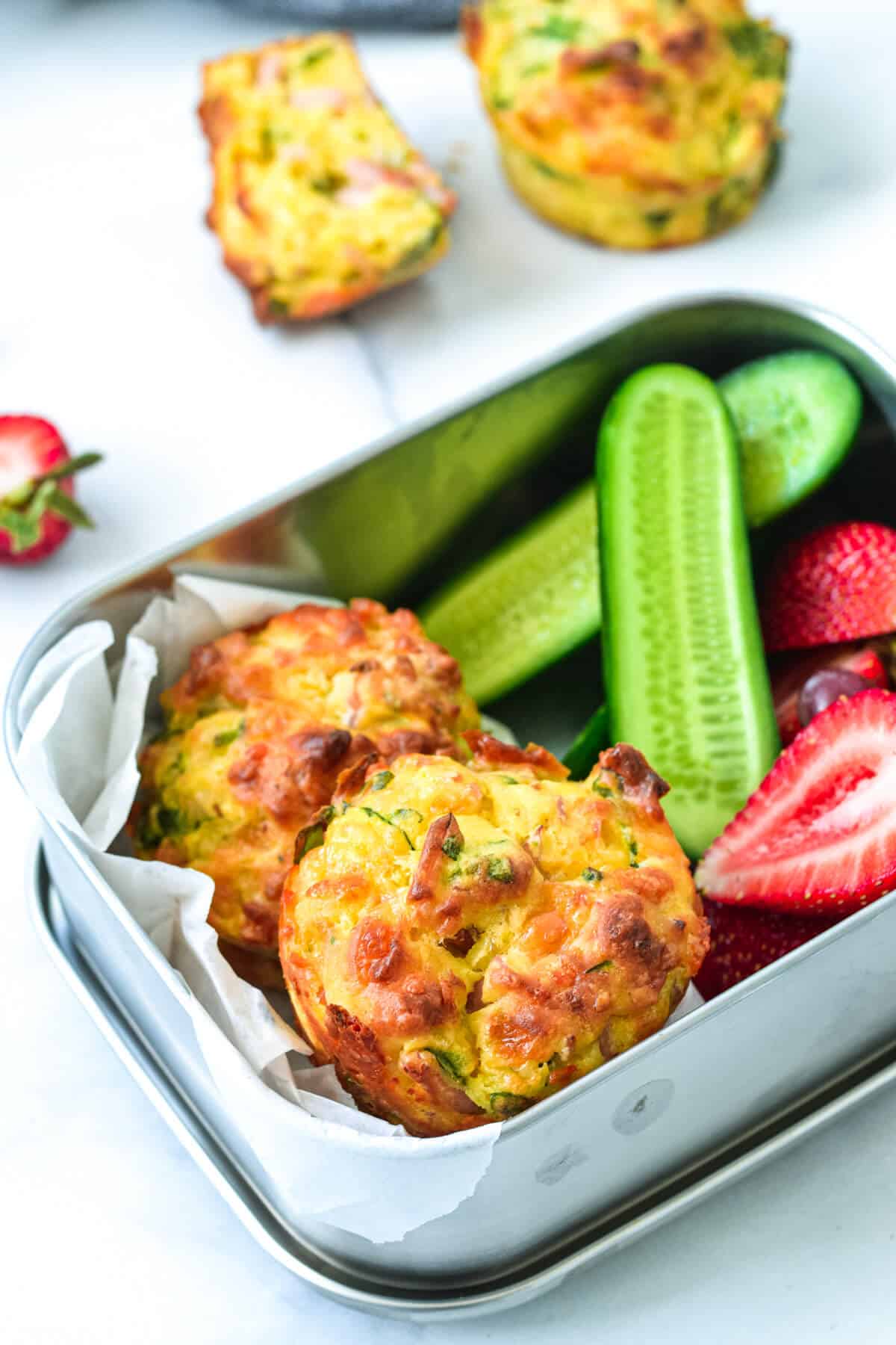 finished vegetable muffins in a steel lunchbox with strawberries and sliced cucumber