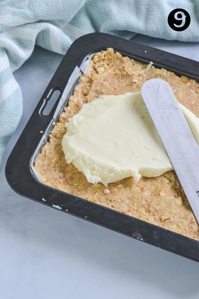 a spatula spreading lemon icing over a biscuit base in a prepared tin