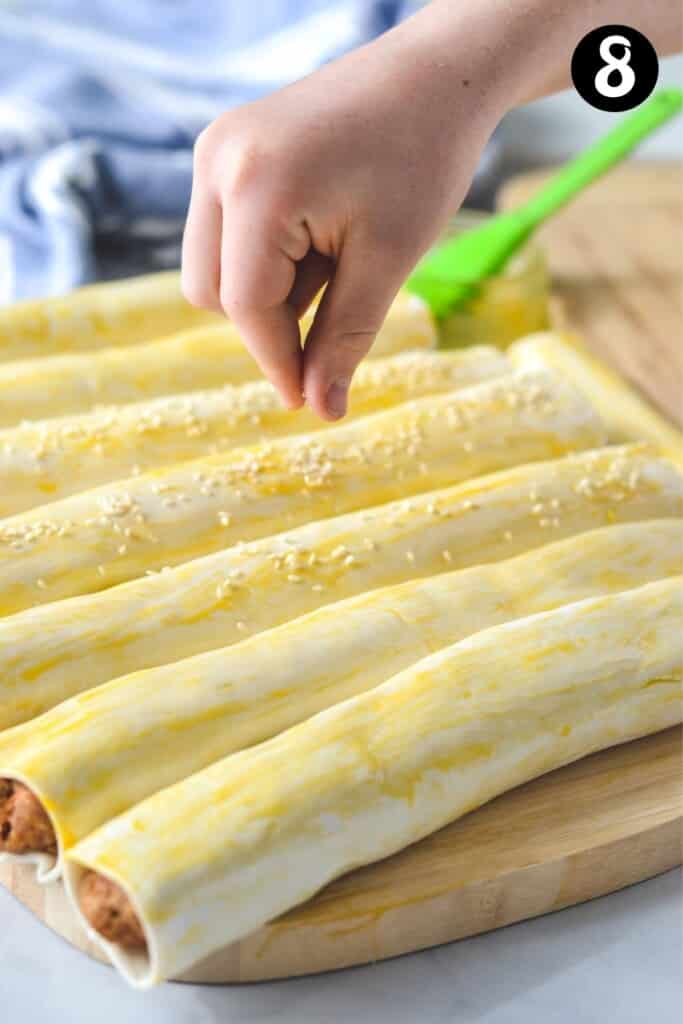 a hand sprinkling sesame seeds over sausage rolls