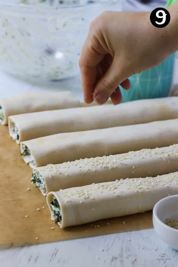 a hand sprinkling sesame seeds over spinach and ricotta rolls