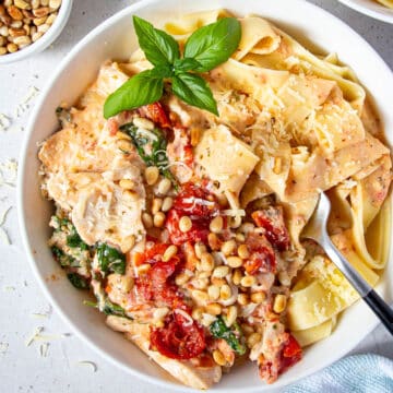 a bowl of pasta topped with chicken, sun dried tomatoes, pine nuts and fresh basil leaves.