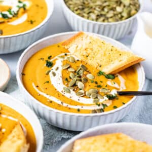 a bowl of pumpkin and carrot soup topped with cream, seeds and bread.