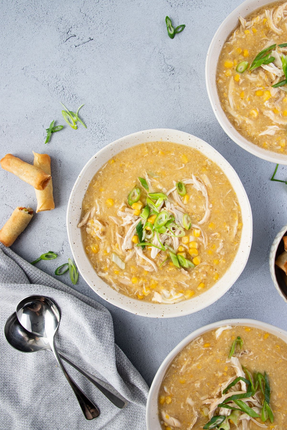 bowls of chicken and corn soup on a grey table topped with slices of spring onion.