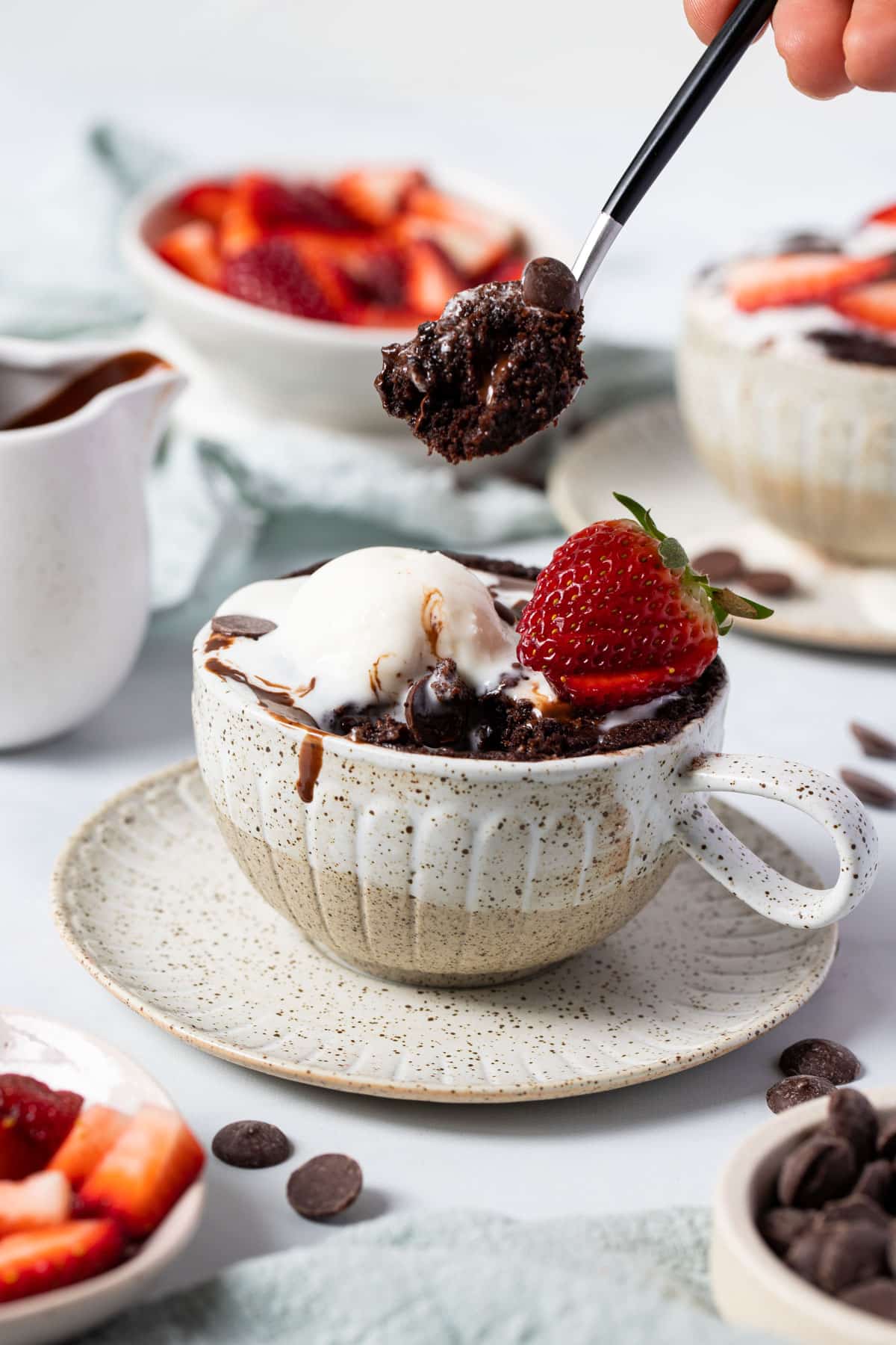 a hand holding a spoon with chocolate cake and a mug of cake on a table.