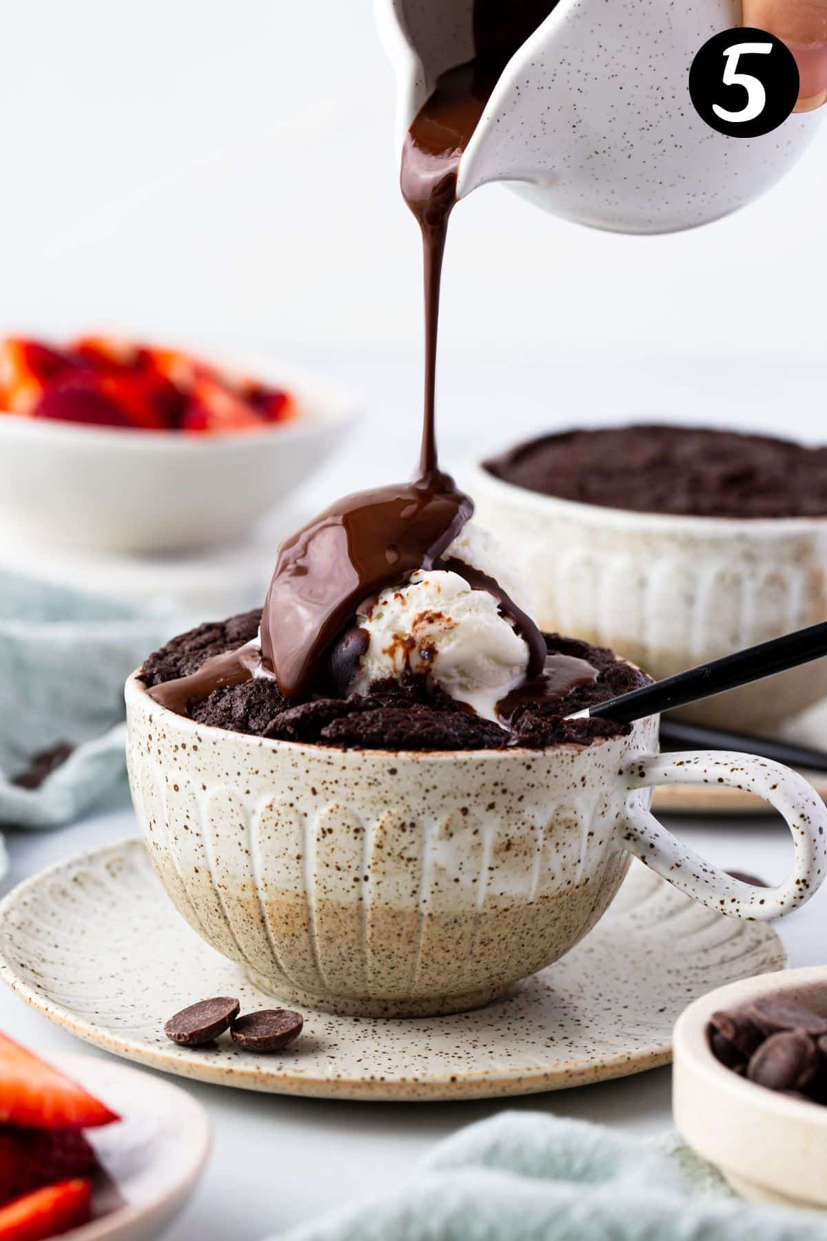 a hand pouring chocolate sauce over a chocolate mug cake topped with ice cream.