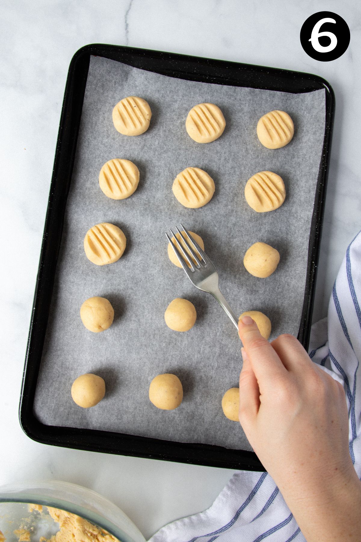 a hand using a fork to flatten balls of dough on a baking tray.