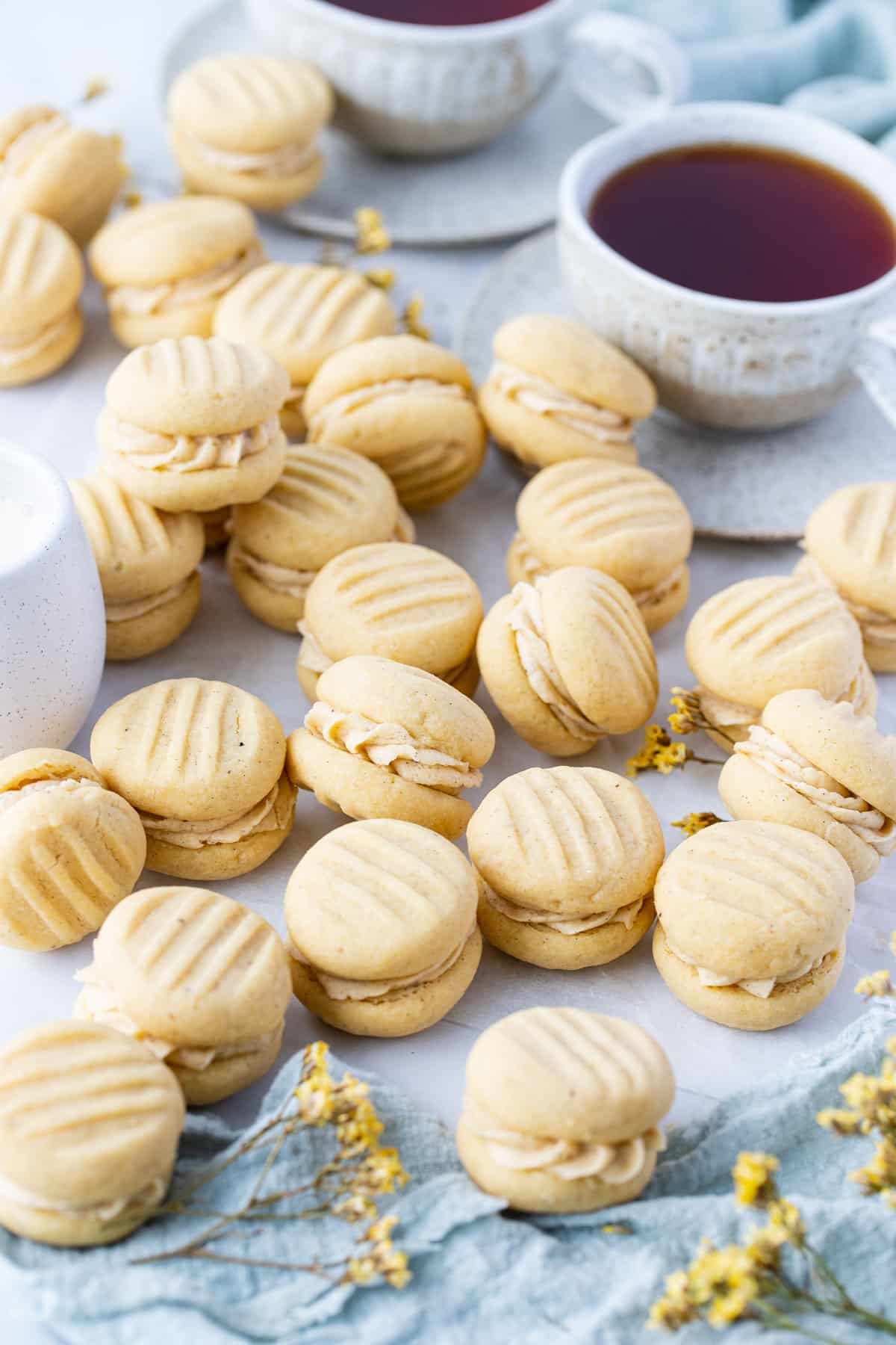 melting moments biscuits filled with vanilla buttercream, spread out on a table with cups of tea.