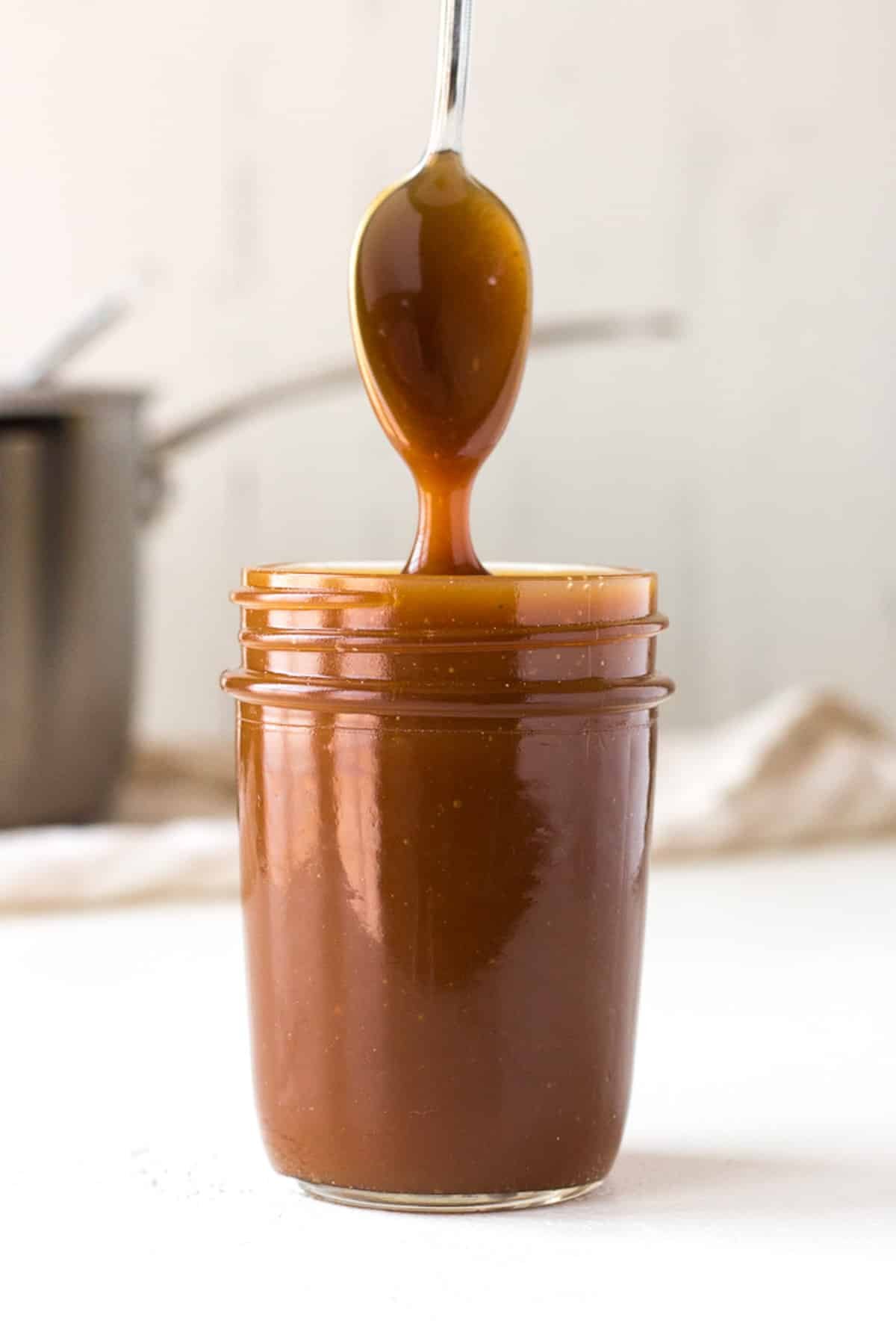 a spoon being held over a jar of caramel sauce, with sauce dripping from the spoon.