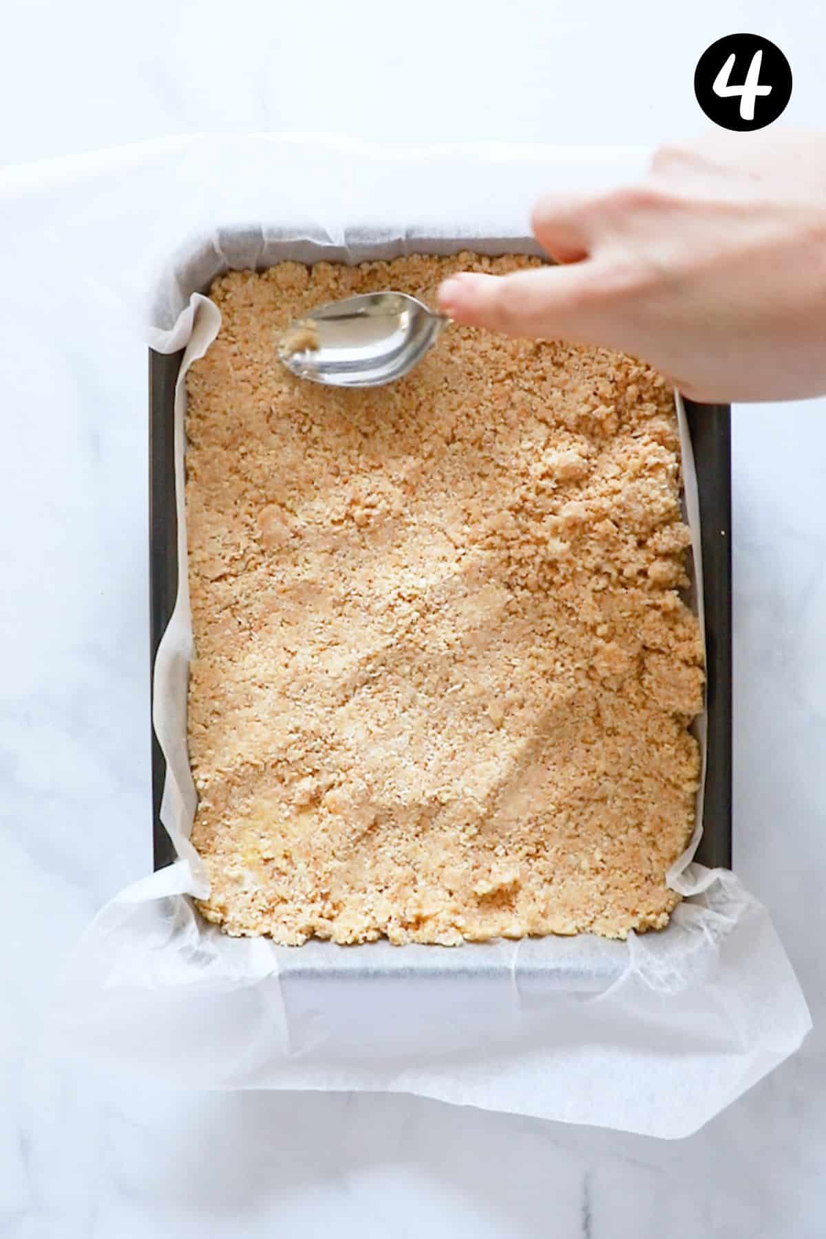a hand pressing a spoon onto the top of slice mixture in a baking tin.