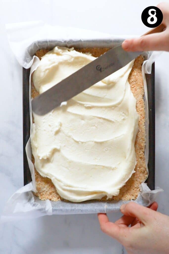 a hand using a spatula to spread icing over a biscuit base pressed into a baking tin.