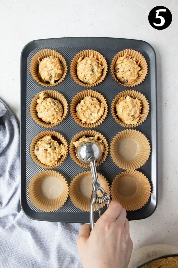 a hand using an ice cream scoop to add muffin batter to a muffin tray.
