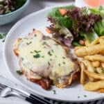 chicken parmigiana topped with ham on a plate with chips and salad.