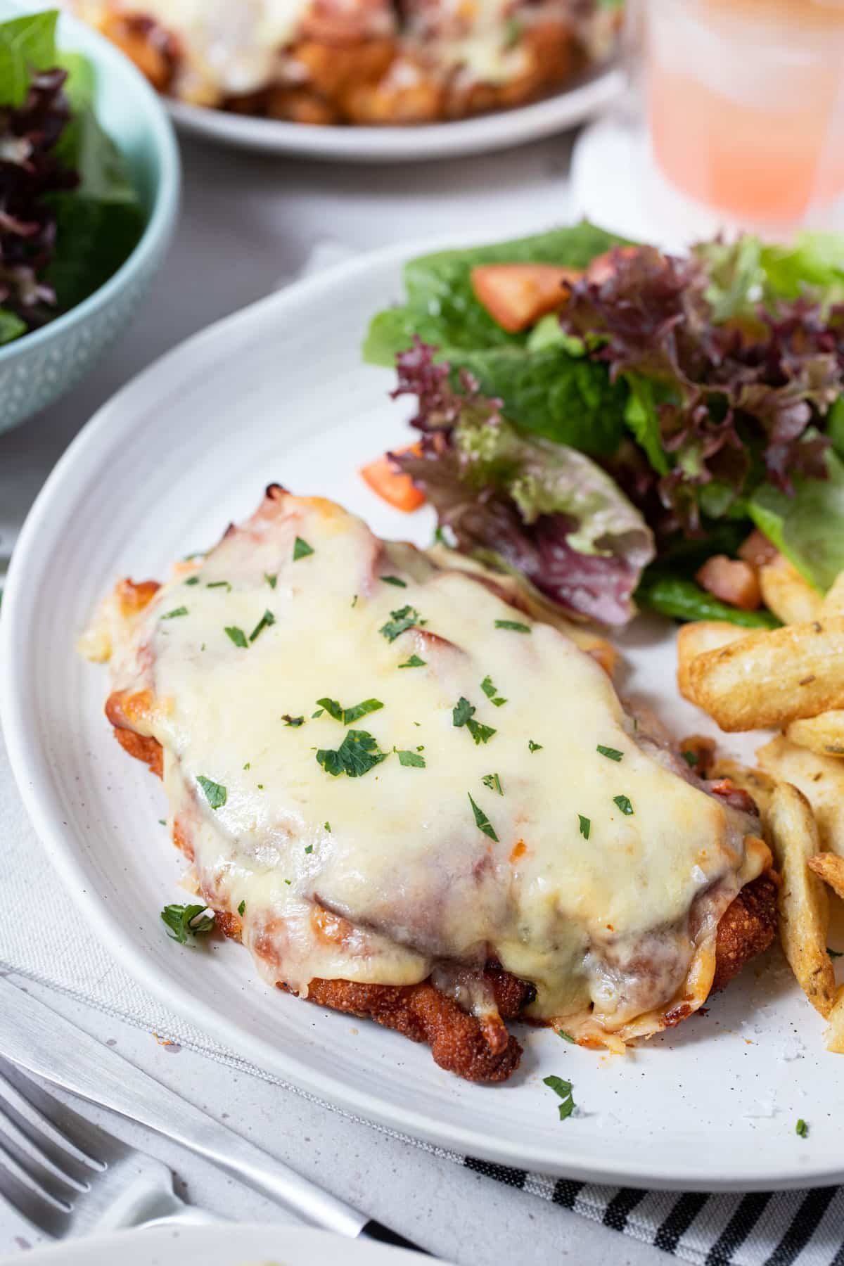 chicken parmigiana topped with ham and cheese on a plate with chips and salad.