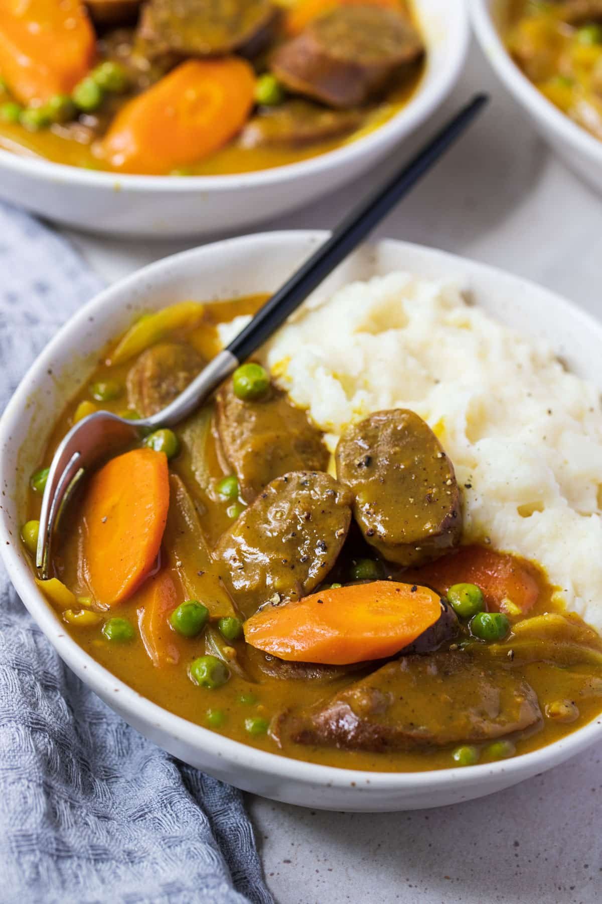 a white bowl on a table filled with sausages in gravy with vegetables and mashed potatoes.