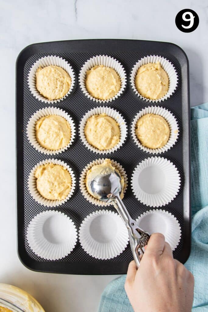 a hand holding an ice cream scoop placing cupcake batter in a baking tray.