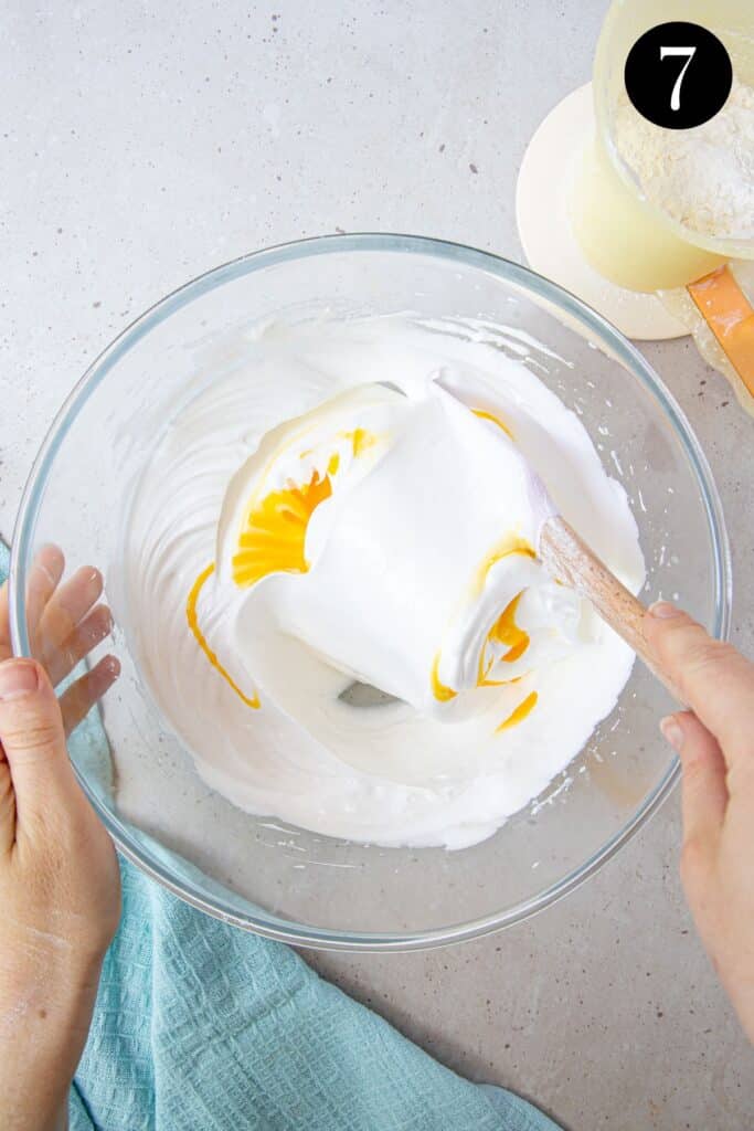 hands folding egg yolks into egg white mixture in a bowl.