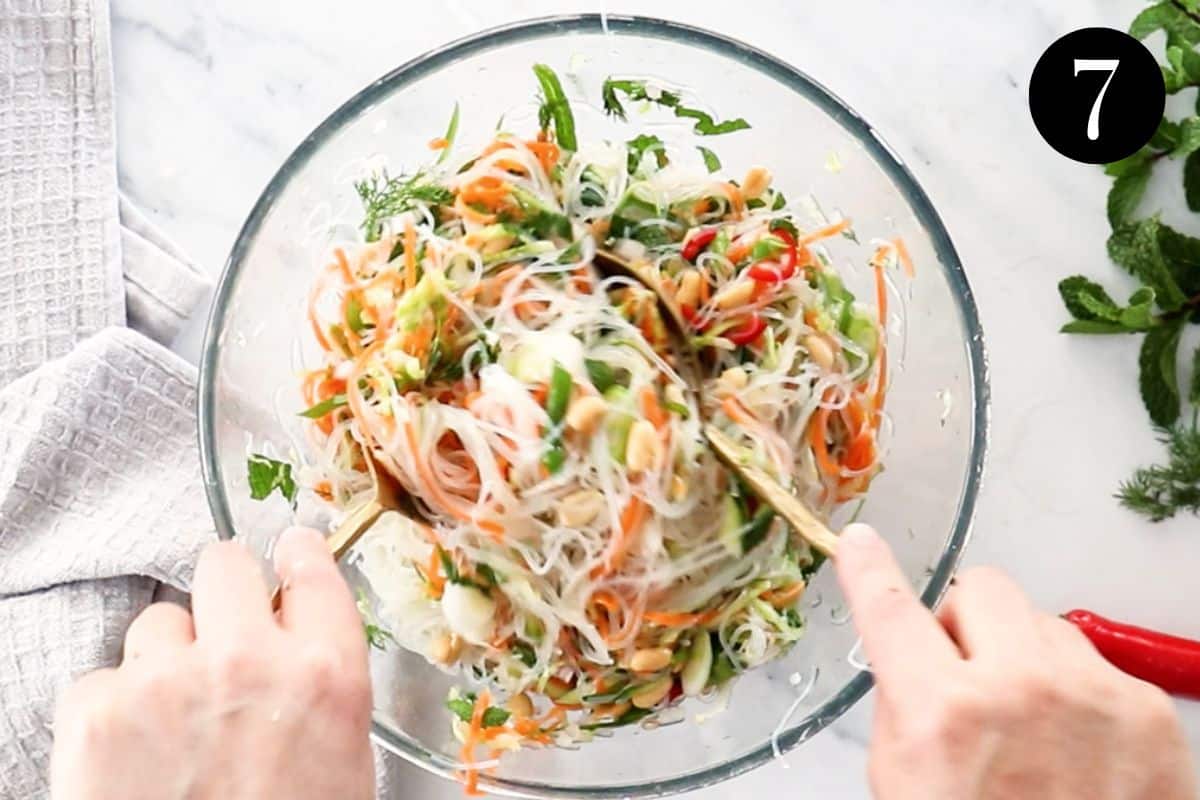 hands stirring a bowl of noodle salad with vegetables.