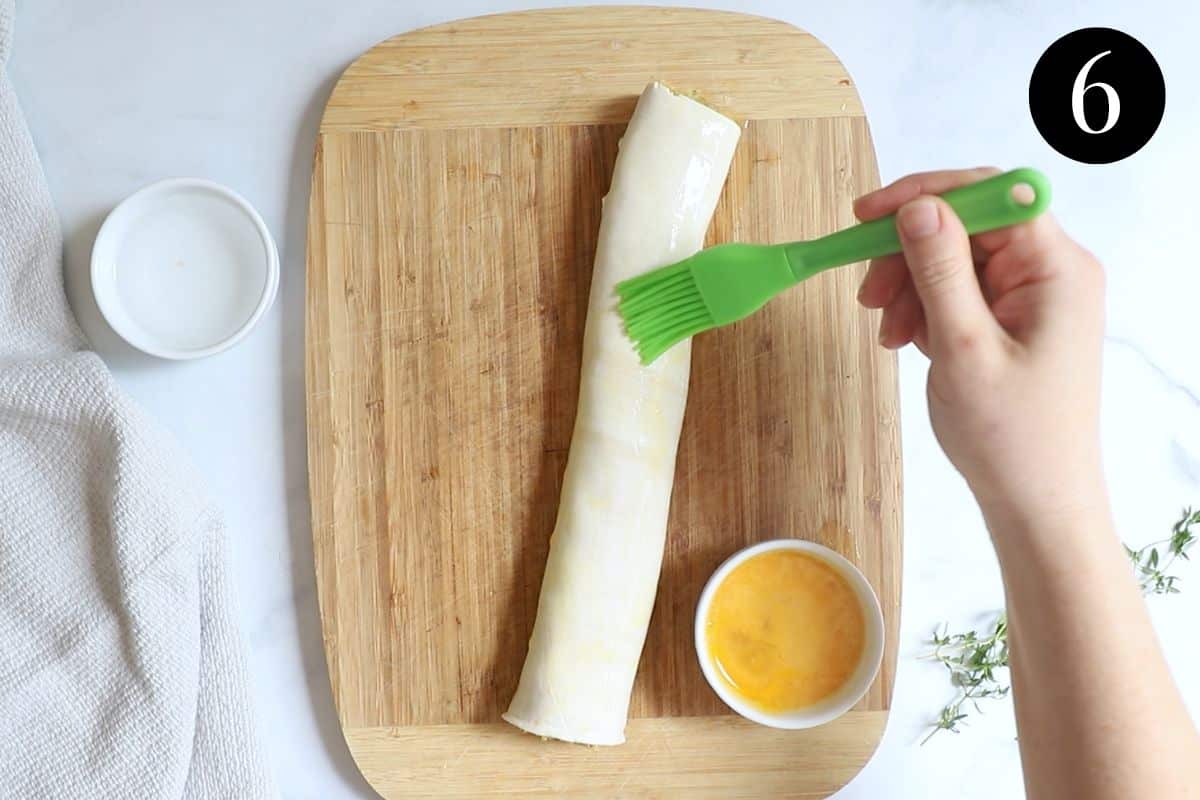 hands using a pastry brush to brush egg over the top of a sausage roll.