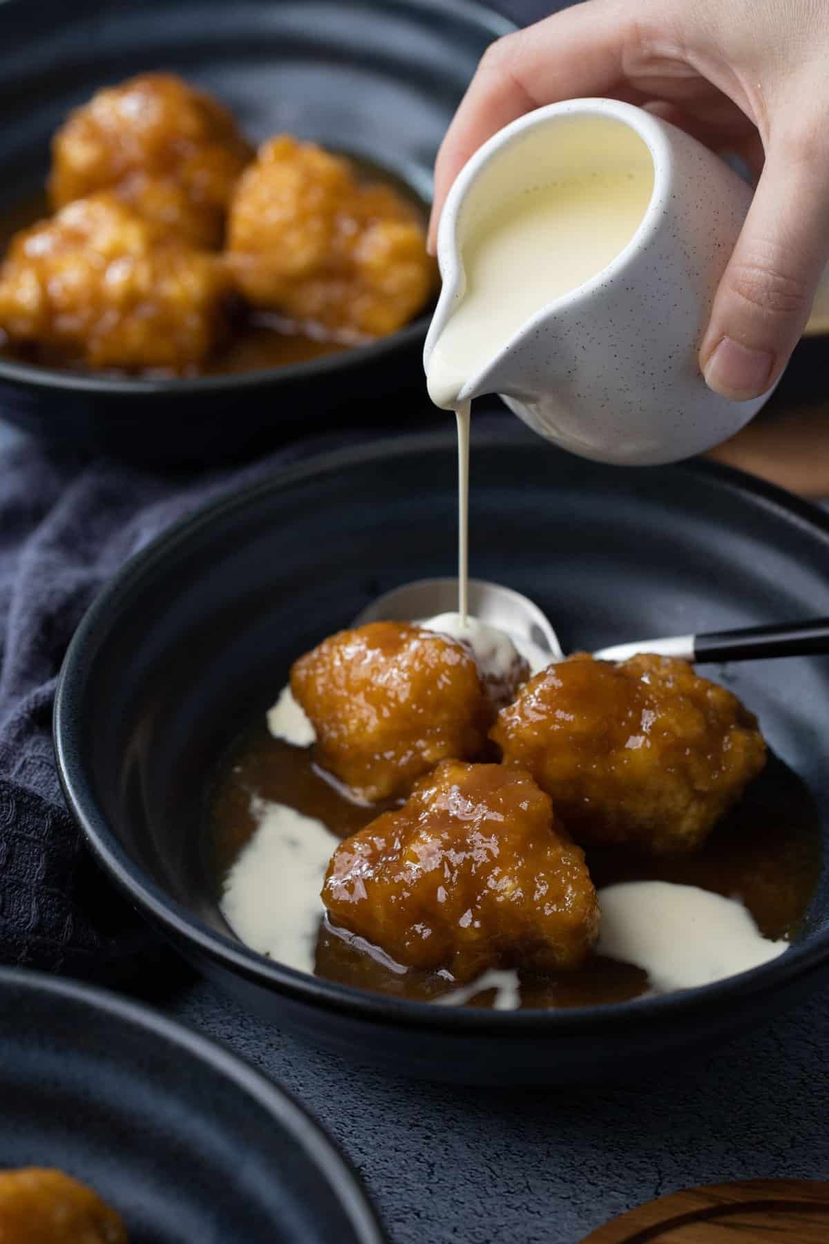 a hand pouring a jug of cream over golden syrup dumplings in a bowl.