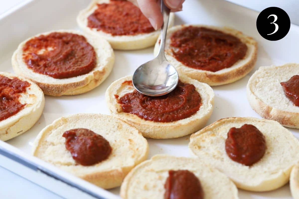 a spoon spreading pizza sauce over English muffins.