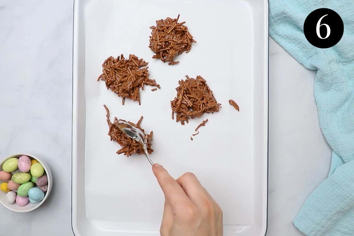 a hand using a spoon to shape chocolate nests on a baking tray.