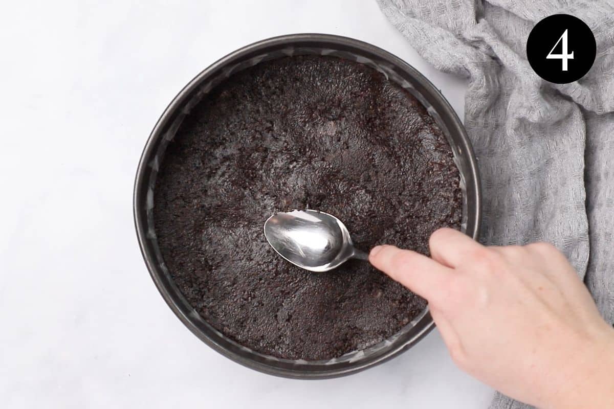 a hand using a spoon to press a biscuit base into a round tin.