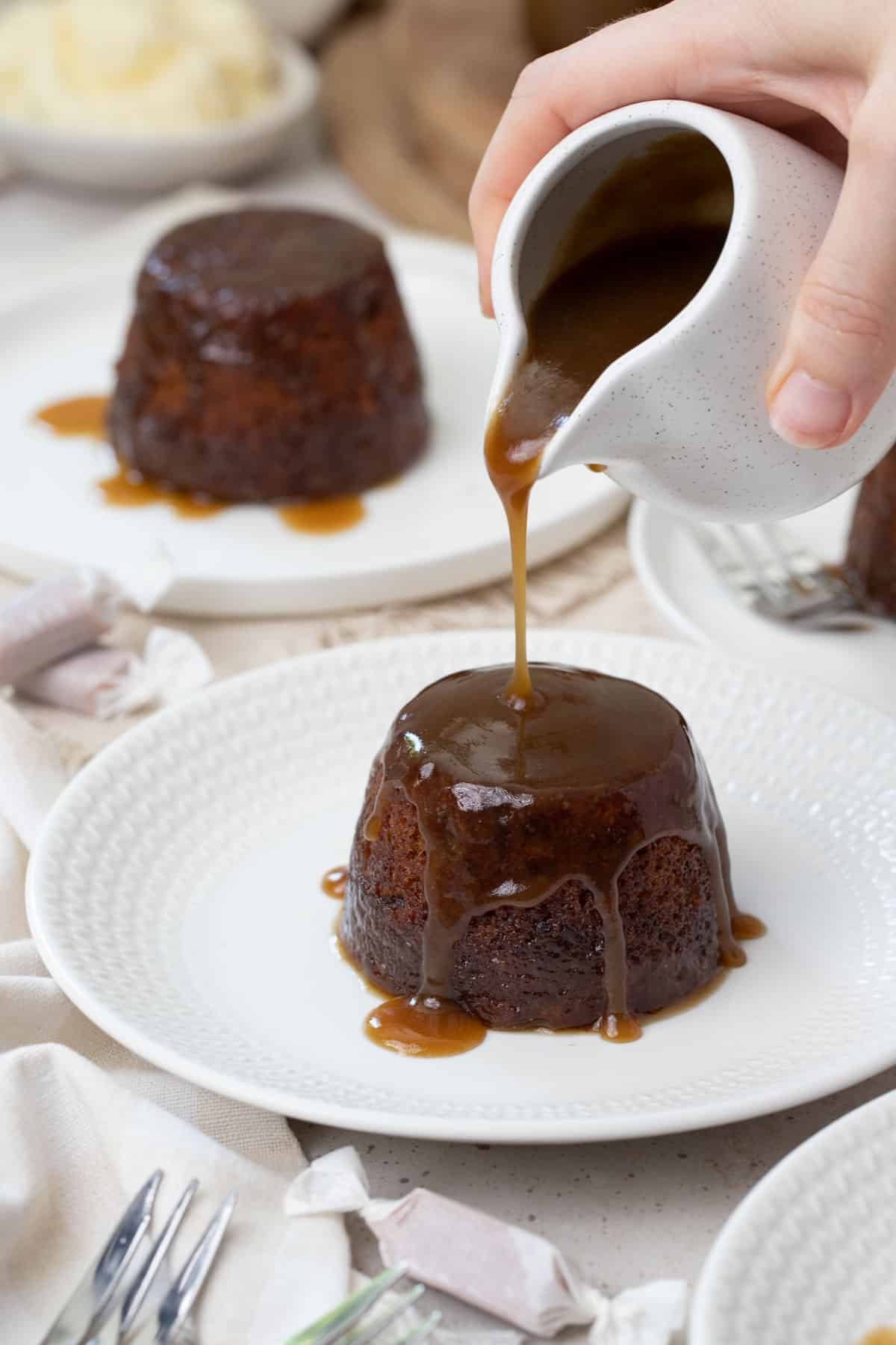 an individual pudding on a white plate. A hand is pouring a jug of caramel sauce over the pudding.