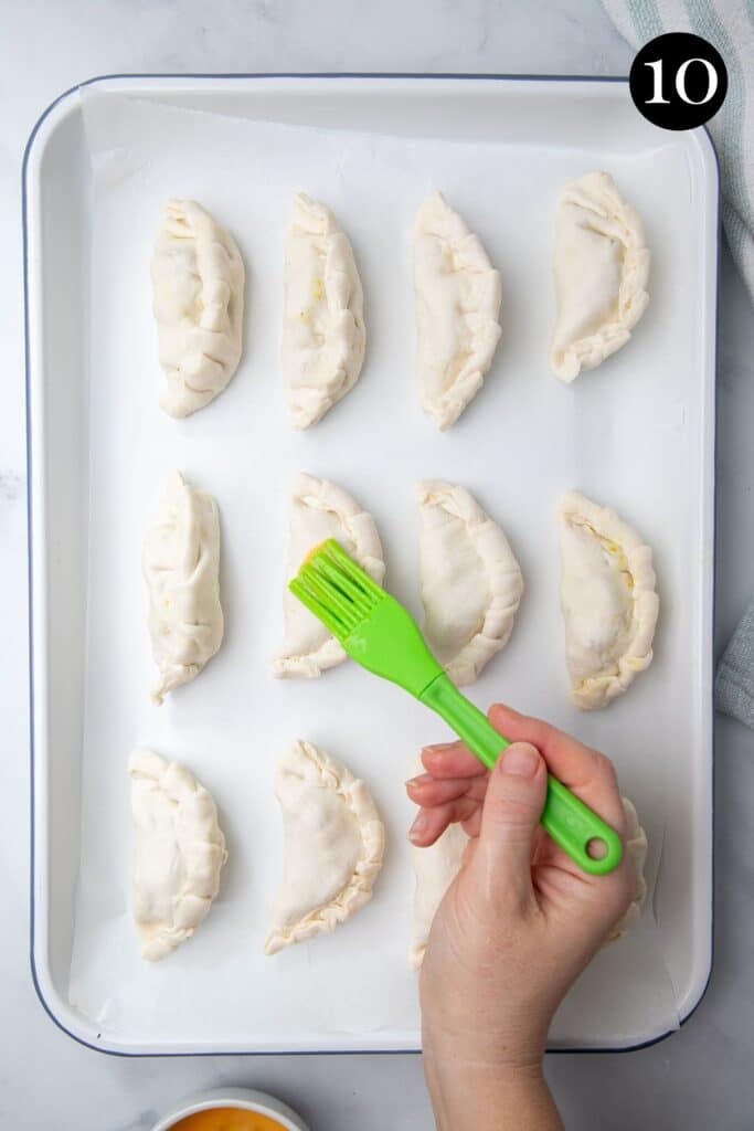pasties on a white tray. A hand is brushing the tops with egg.