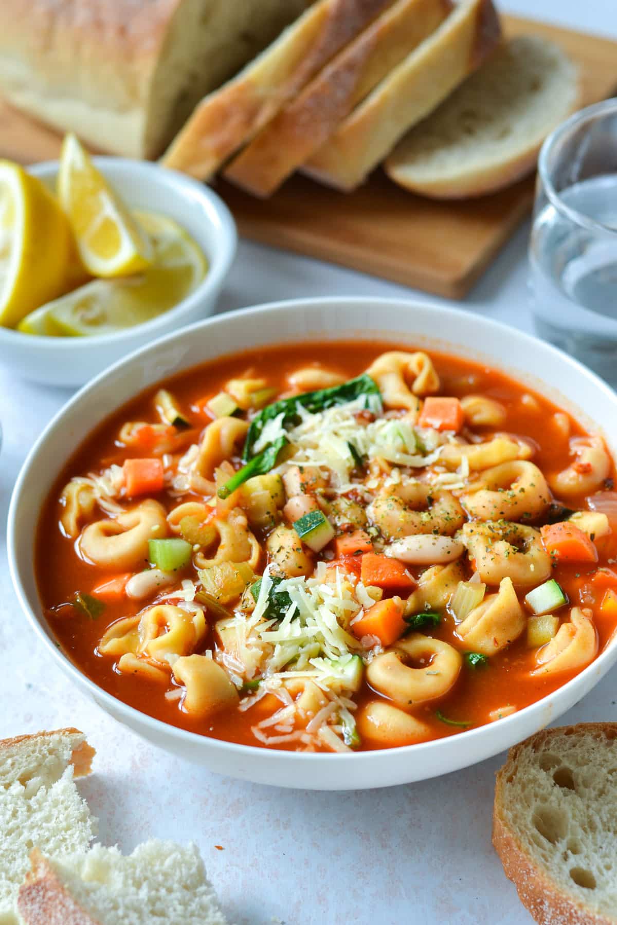 a bowl of tortellini soup on a table with bread and cheese.