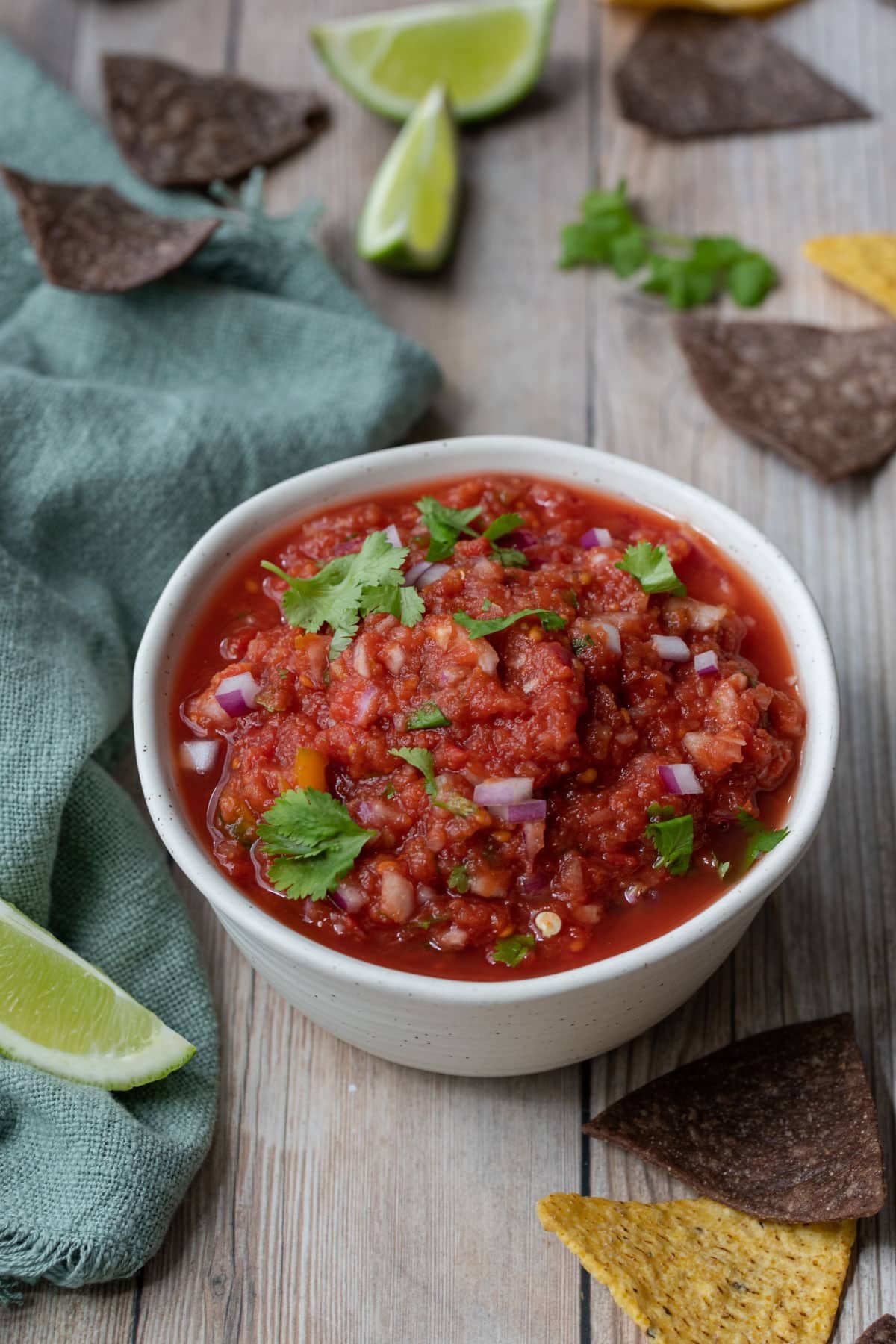 a bowl of tomato salsa on a table with corn chips and lime wedges.