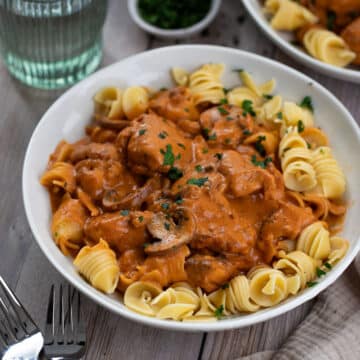 a bowl of chicken stroganoff on a table, with pasta and herbs.