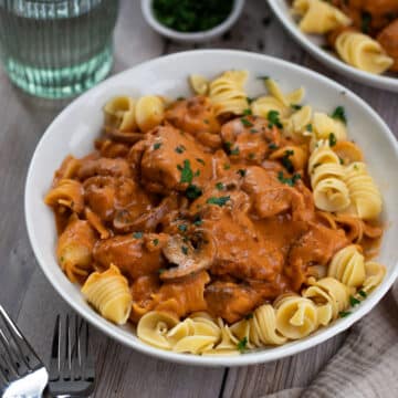 a bowl of chicken stroganoff on a table, with pasta and herbs.