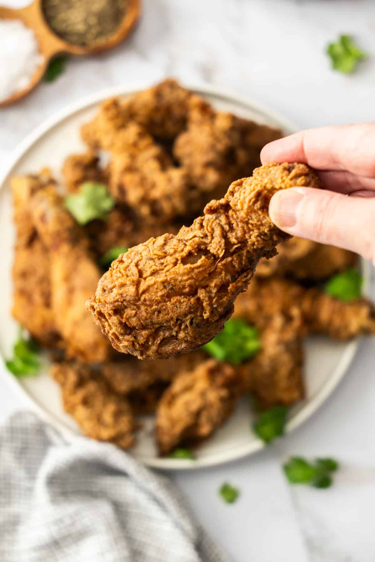 a hand, holding a fried chicken drumstick over a plate of fried chicken.