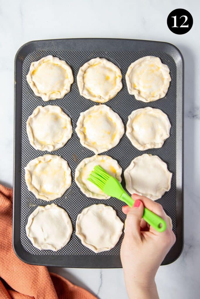 a hand, brushing egg over pies with puff pastry lids.