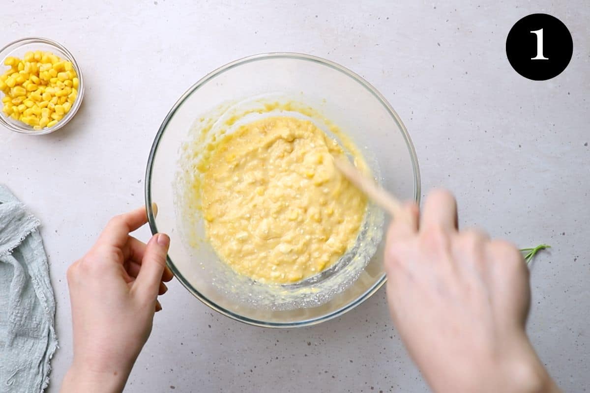 hands using a wooden spoon to mix a bowl of creamed corn and wet ingredients.