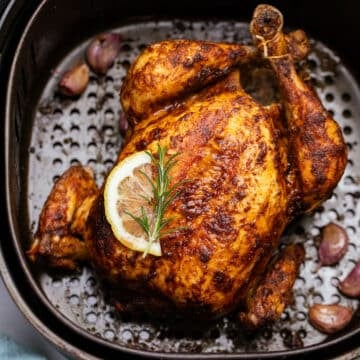 a whole chicken in an air fryer basket, topped with herbs and a slice of lemon.