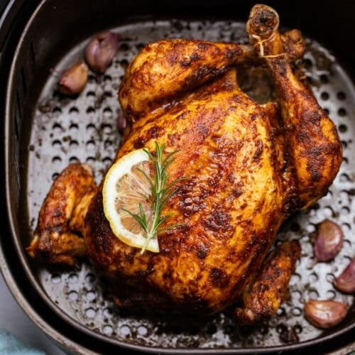 a whole chicken in an air fryer basket, topped with herbs and a slice of lemon.
