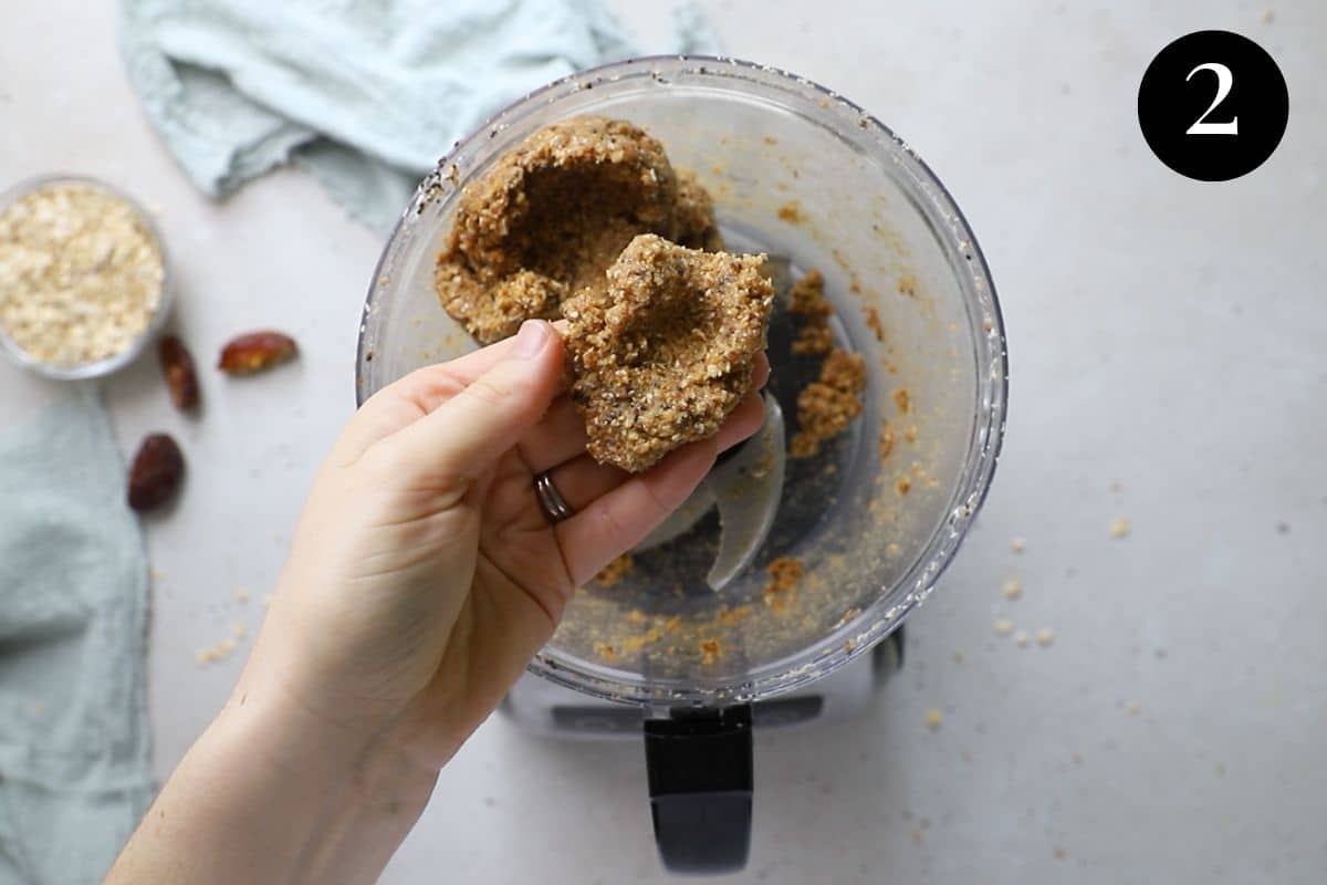 a hand holding a thick bliss ball mixture above a food processor.