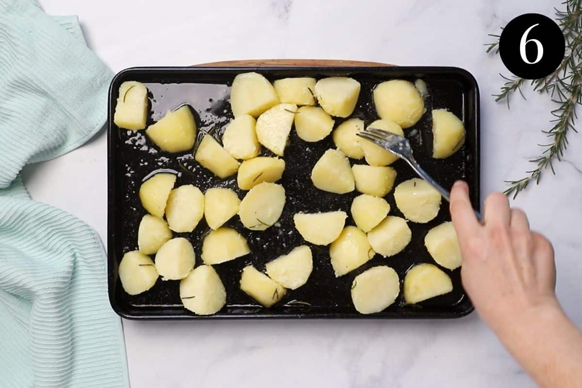 A roasting tray with potato pieces and duck fat. A fork is being used to turn the potatoes in the hot duck fat.