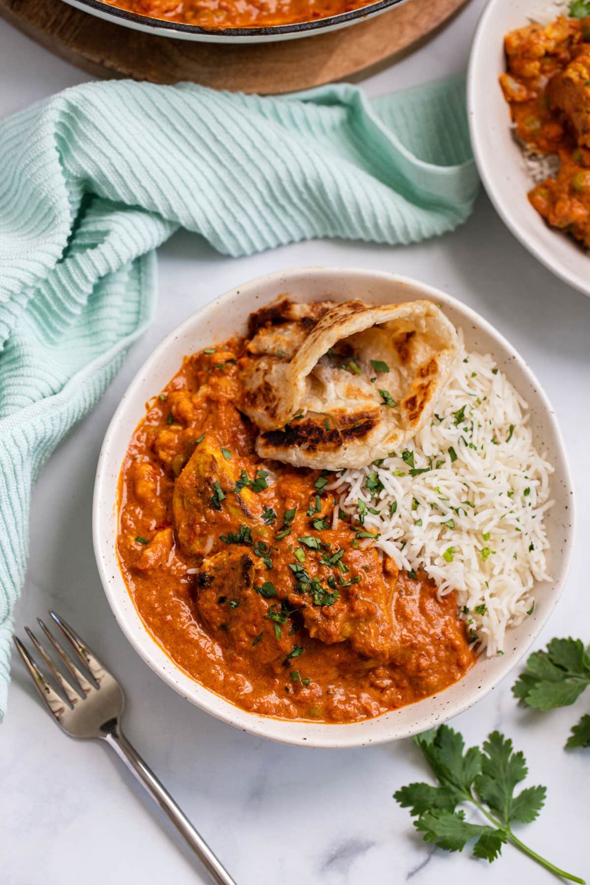 a bowl of tikka masala on a table with naan bread and herbs.