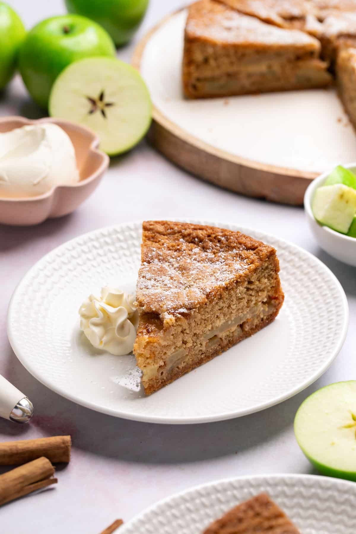 a slice of cake on a table with sliced apples and cream.