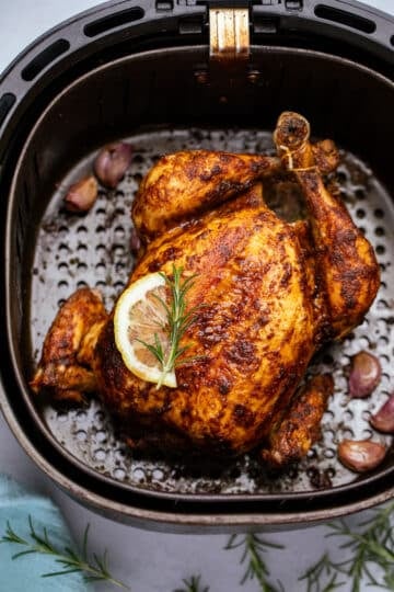 a whole roast chicken in an air fryer basket. It is topped with a slice of lemon and herbs.