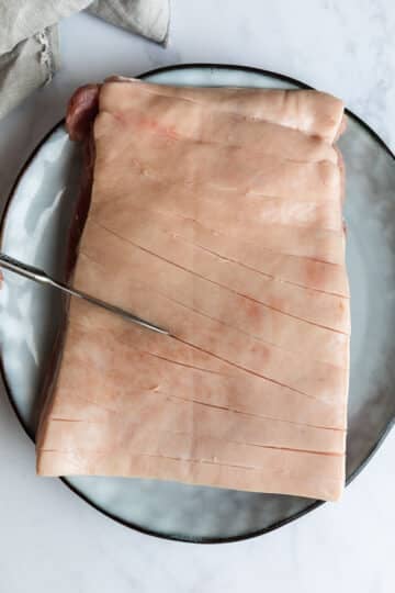 pork belly on a plate. A knife is scoring the skin on top.
